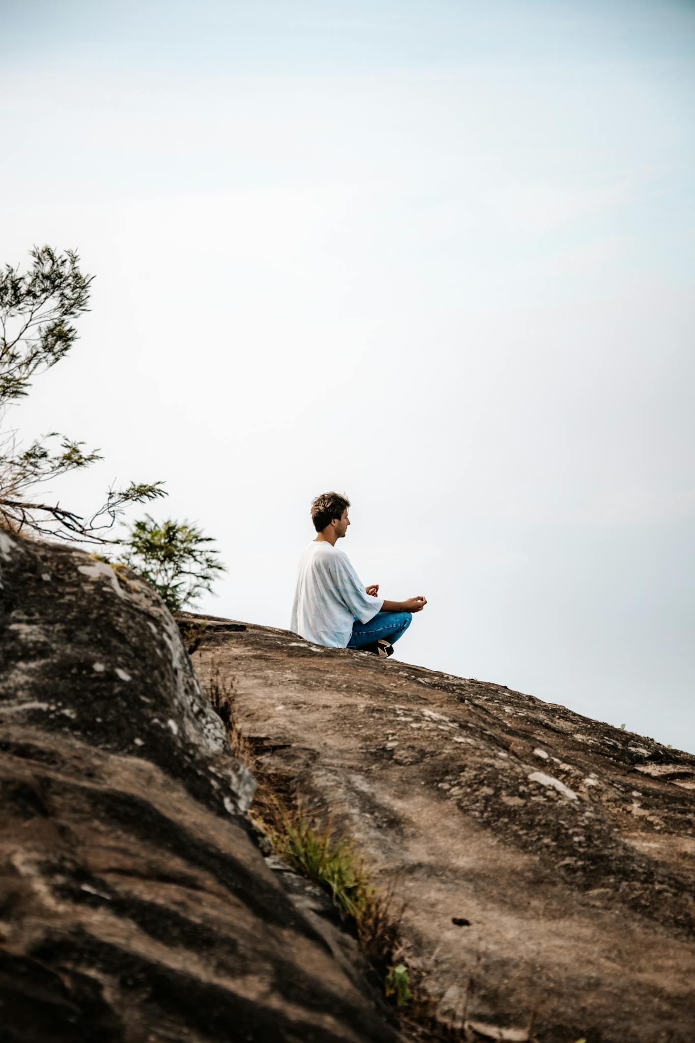A man sits meditating on a rocky hill under clear skies, promoting mindfulness and relaxation.